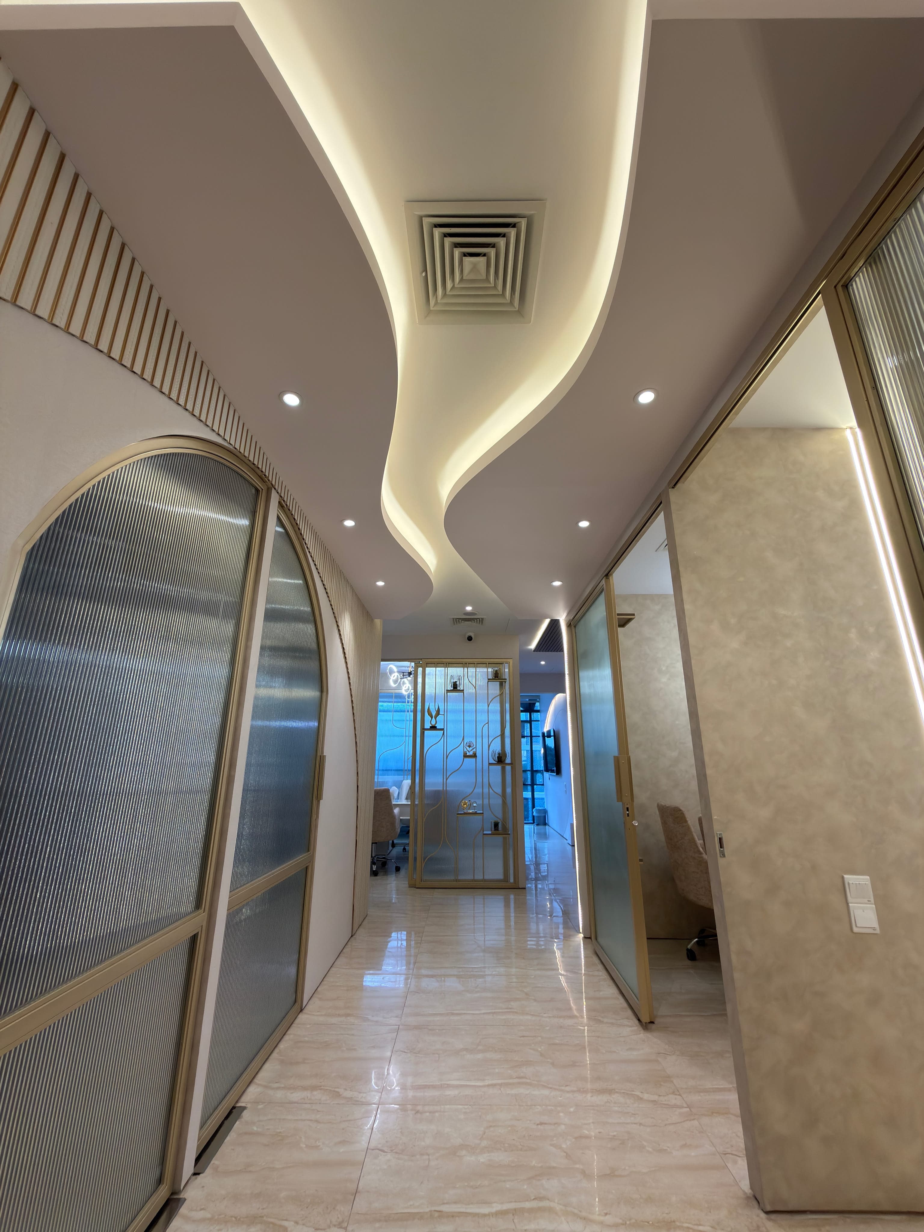 An aesthetic center's corridor: arched brass-framed glass doors and a sculpted cove ceiling lead past a fluted display niche to a marble-floored suite beyond.